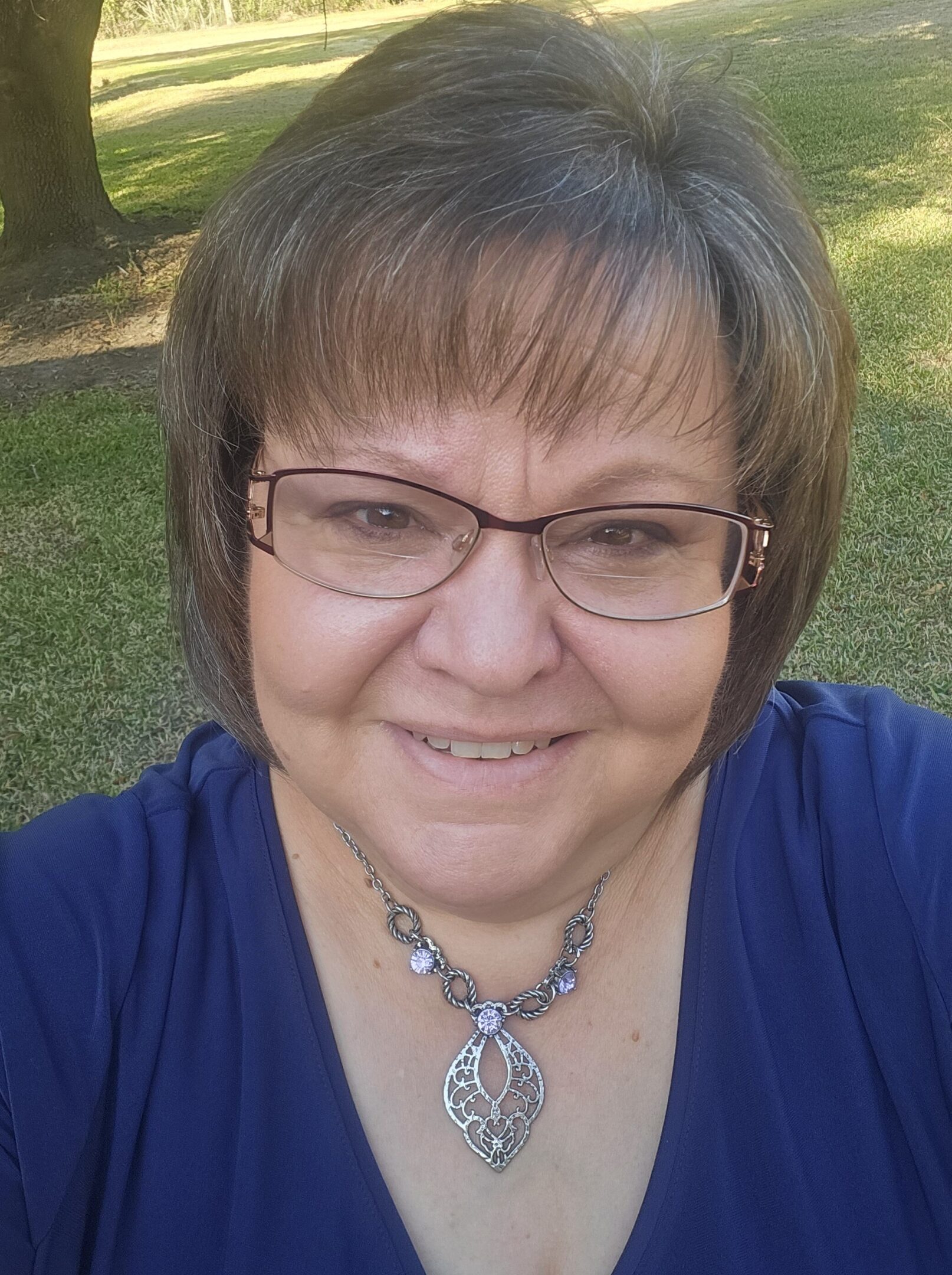 A smiling woman with glasses outdoors, wearing a blue top and a pendant necklace.